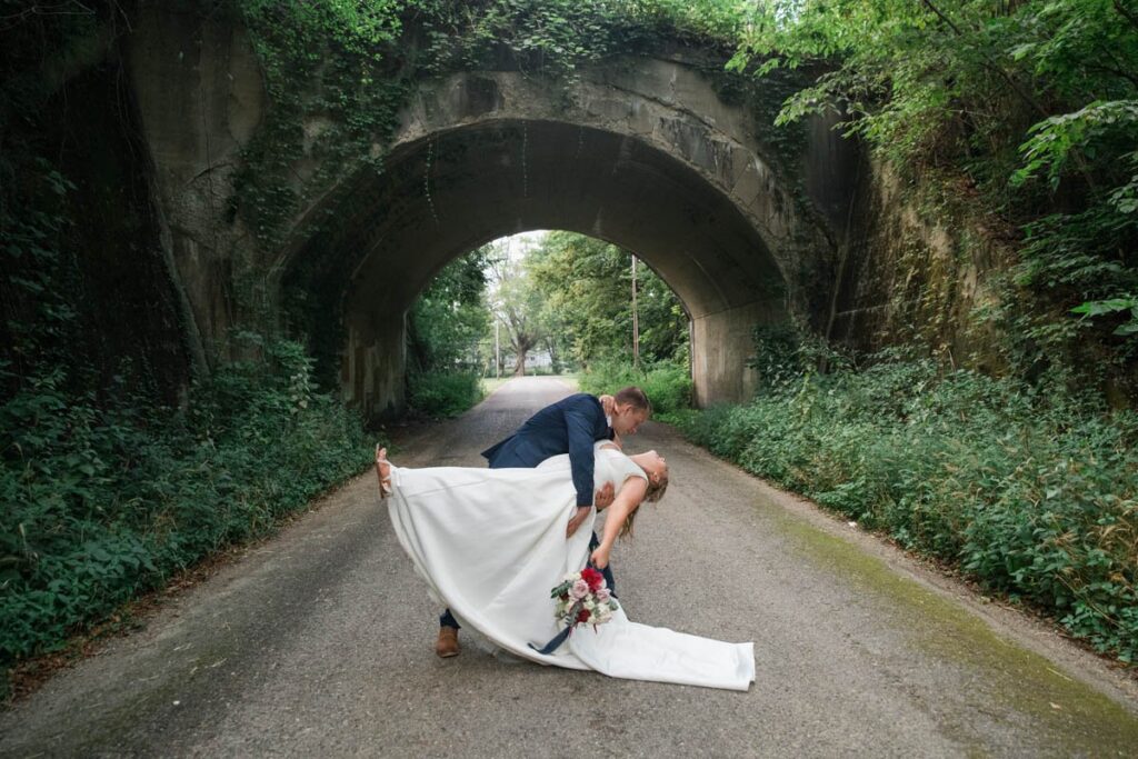 Groom dips bride in front of an old concrete bridge.