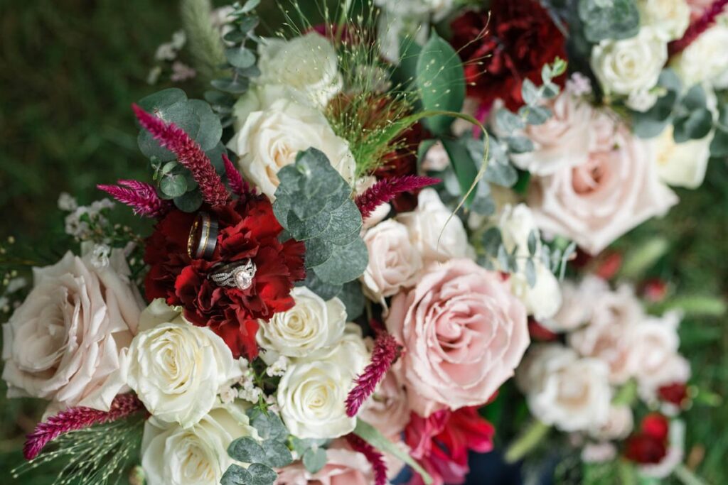 Blush, red, while, and greenery bouquet with wedding rings sitting on flowers.