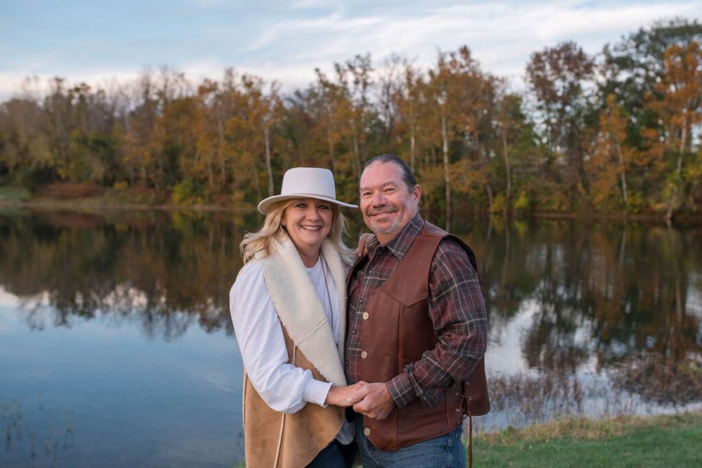 Couple holds one another closely and smiles while standing in front of lake in the fall.