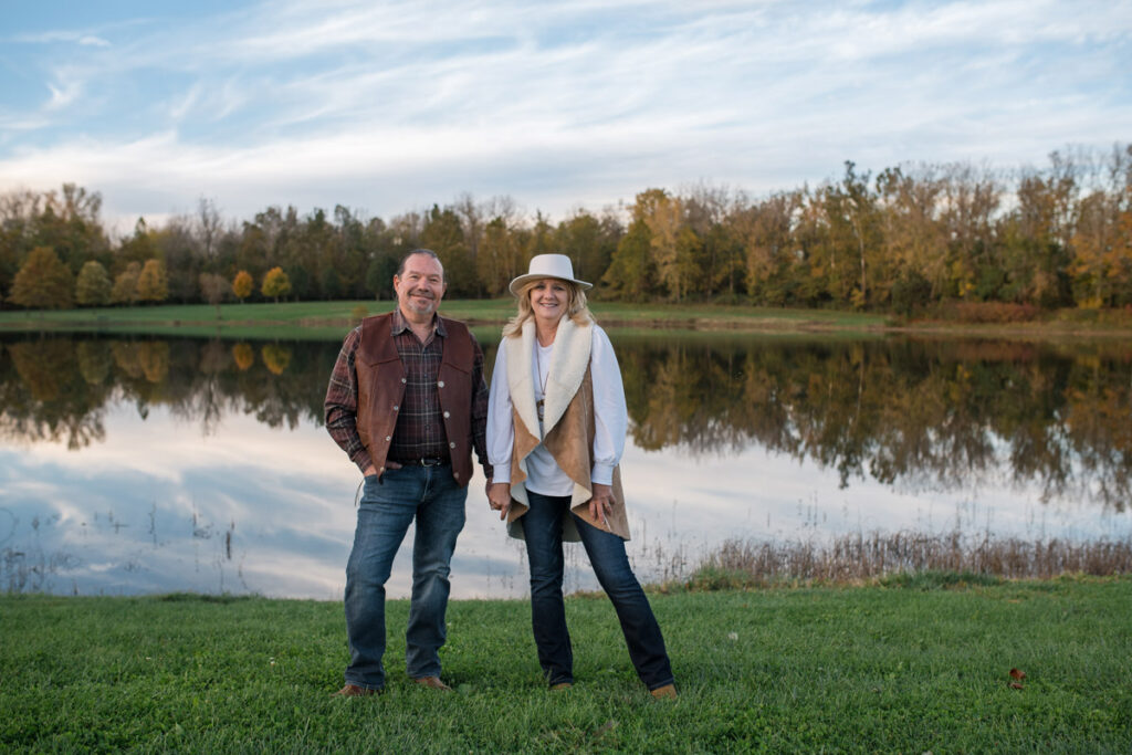 Husband and wife hold hands and smile as they stand in front of Blue Heron Lake.