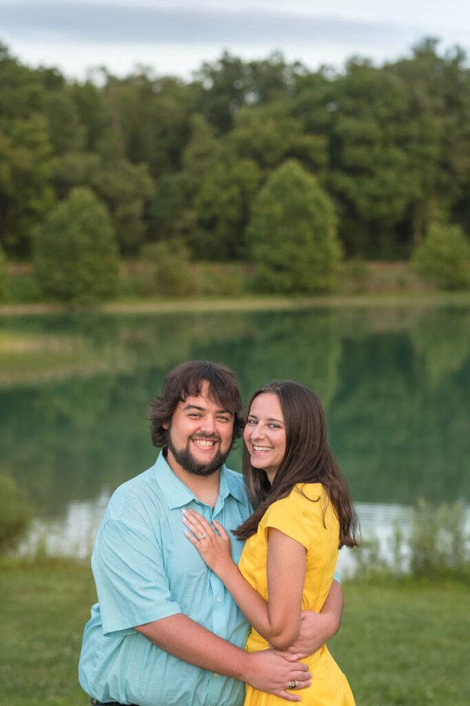 Couple is laughing with arms around each other in front of a lake.