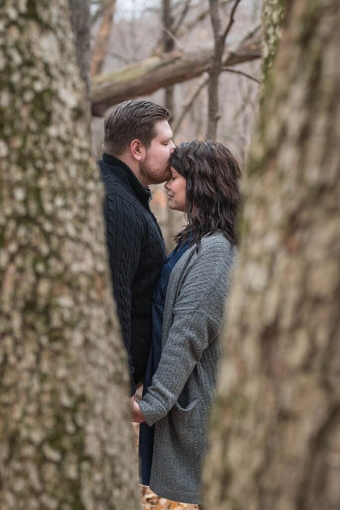Man kisses woman's forehead while they stand amongst the trees.