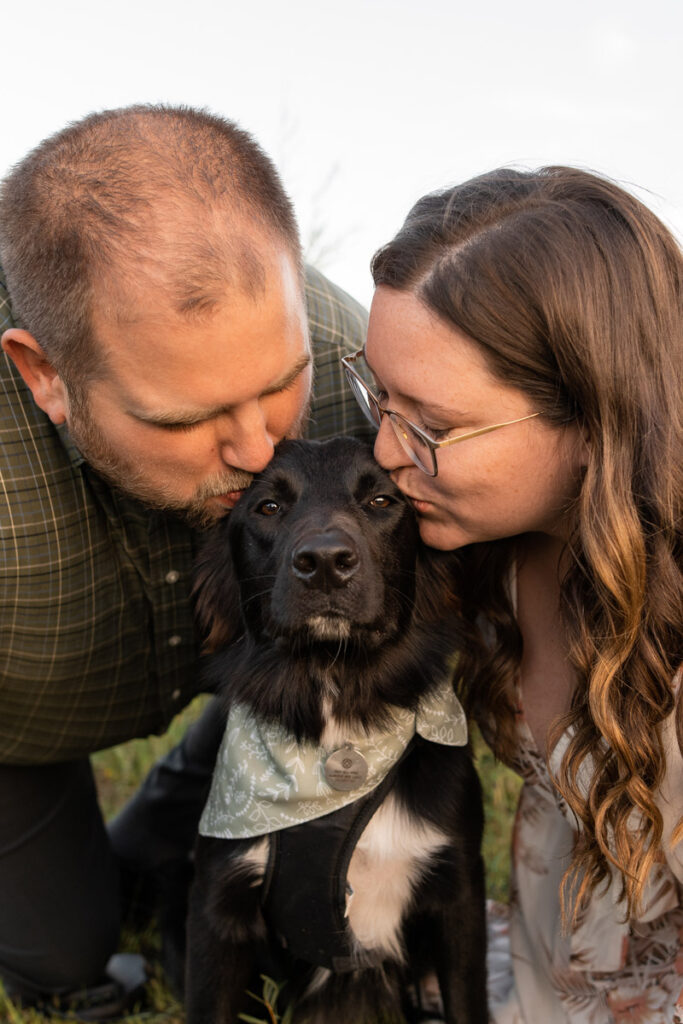 Man and woman kiss their dog while he looks straight ahead.