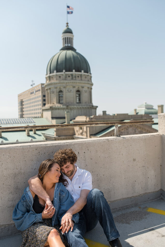 Couple sits with arms around one another on parking garage roof with capital building behind them.