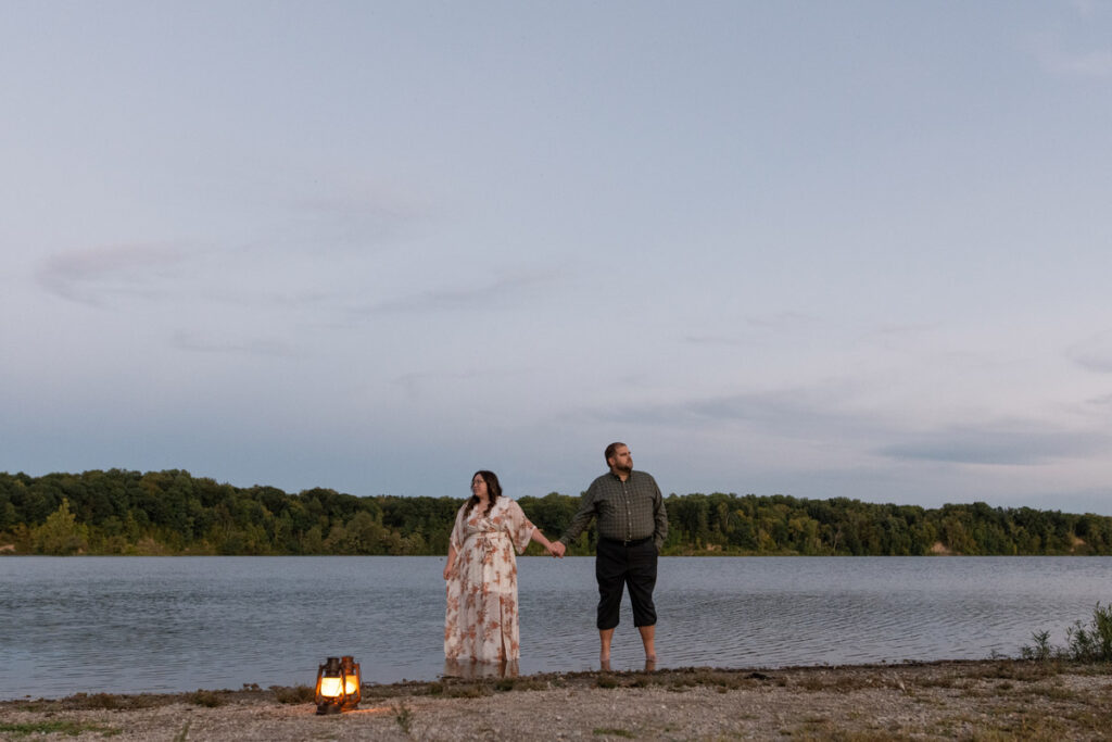 Couple holds hands while standing in lake with lanterns sitting on shore near them.