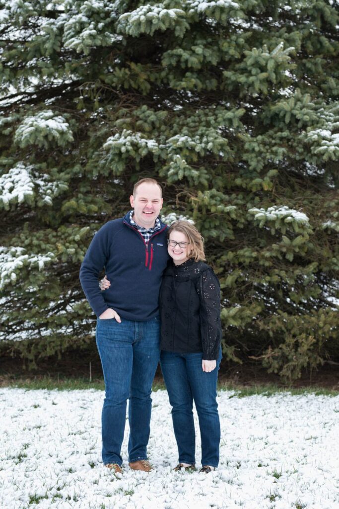 A couple smiles in front of an evergreen tree in the snow.