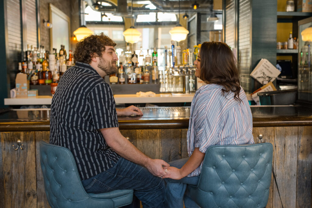 Man and woman hold hands at bar while smiling at one another.