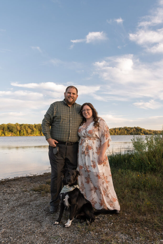 Man and woman stand next to lake and smile with their dog.