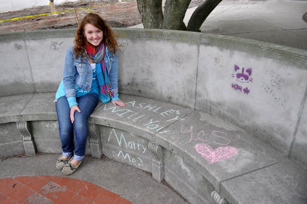 Woman smiles while sitting on a stone bench with "Will you marry me?" written in chalk.