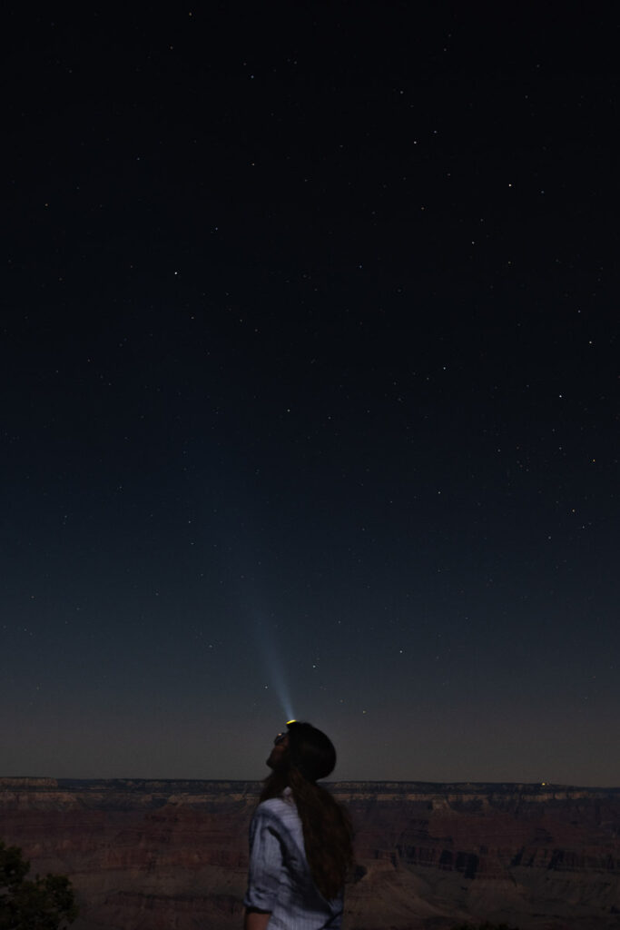 Woman wearing headlamp gazes up at stars above Grand Canyon.