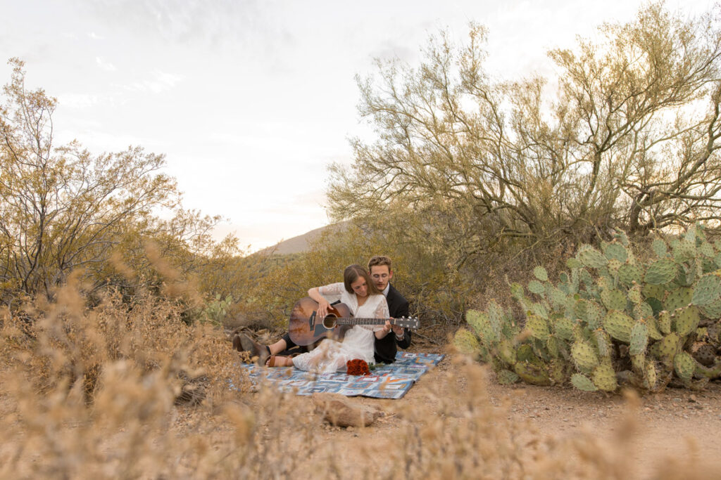 Couple sits on a blanket in the desert playing guitar together on their elopement day.