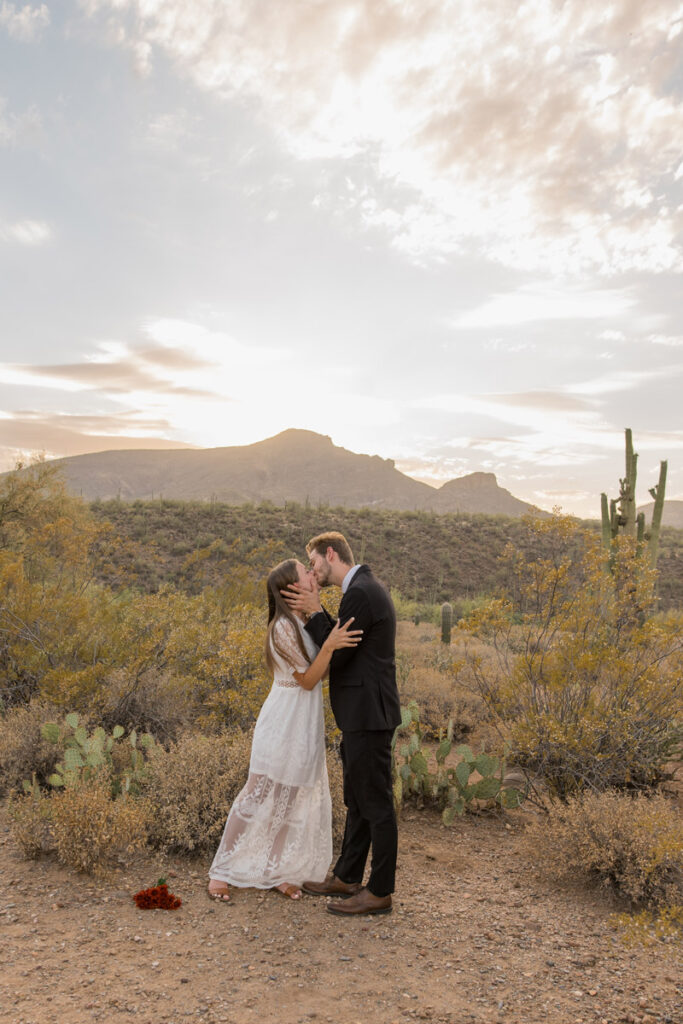 Bride and groom share first kiss during desert sunset.