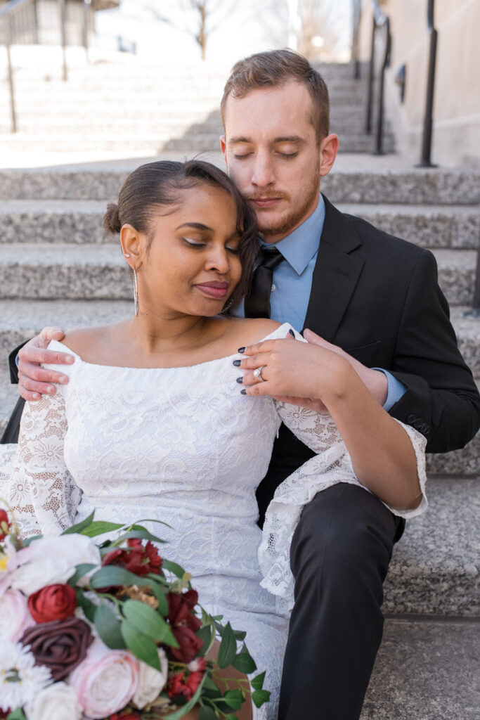 Bride and groom sit on marble steps closely to one another with eyes closed.
