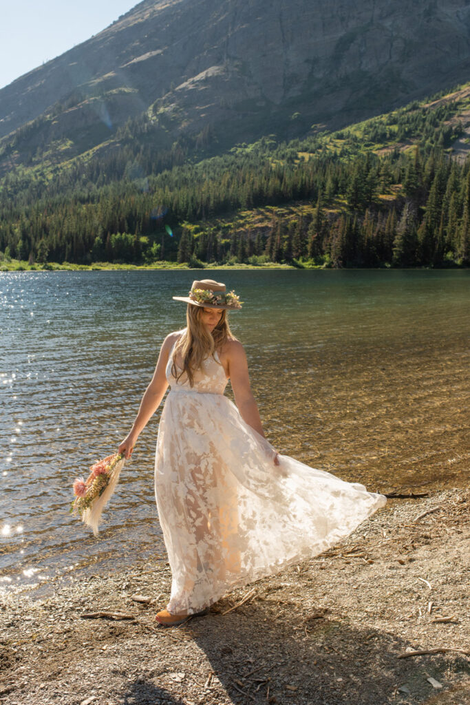 Bride plays with white dress as she walks along a lake shore.