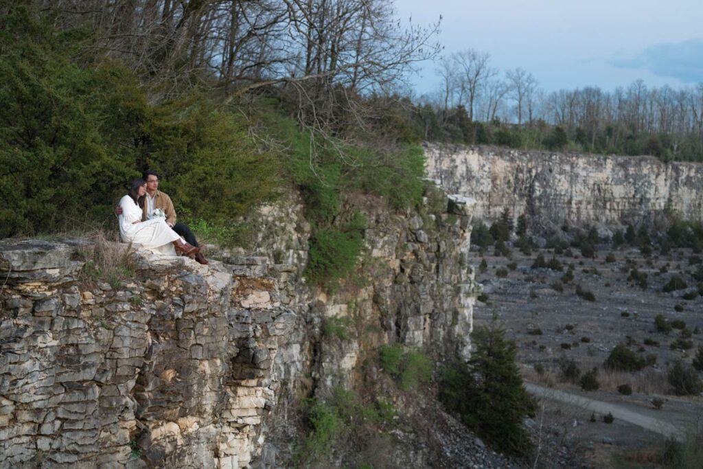 Couple cuddles up on the edge of a cliff looking out into the distance at sunset.