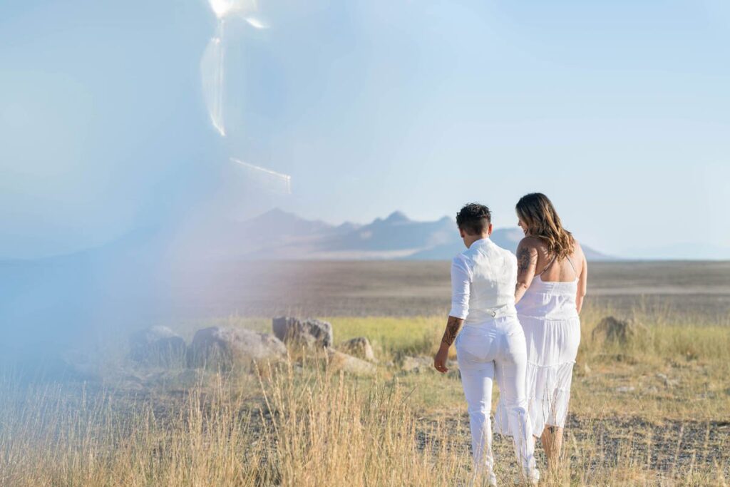 A couple that is wearing white is holding hands and walking in tall grass towards the mountains.