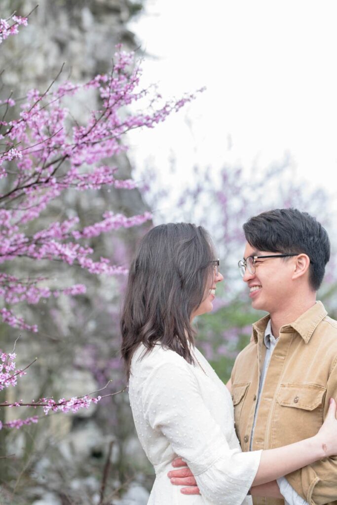 Couple stands amongst purple flowering trees smiling at one another.