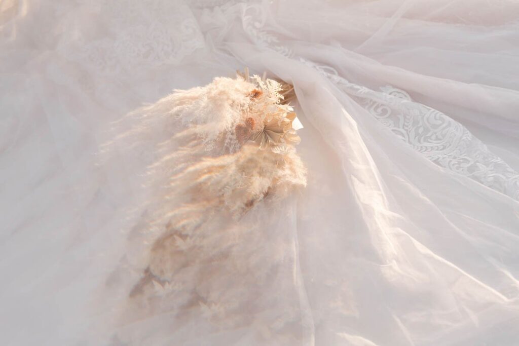 Dried bridal bouquet sits on train on bride's white lace dress.