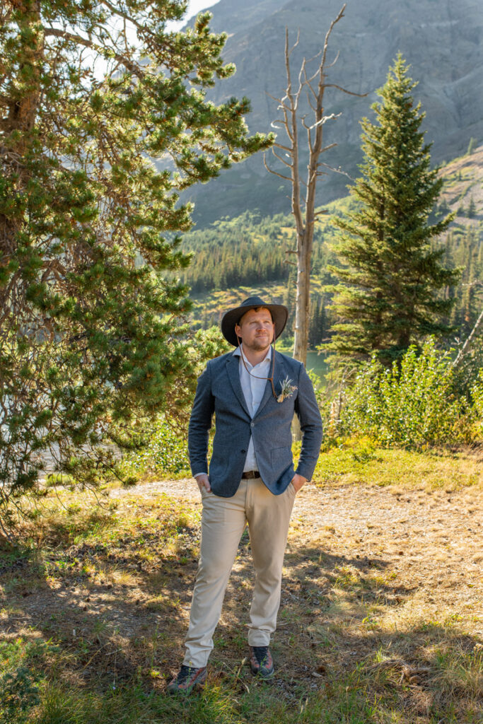 Groom stands by pine trees looking away with hands in pockets.
