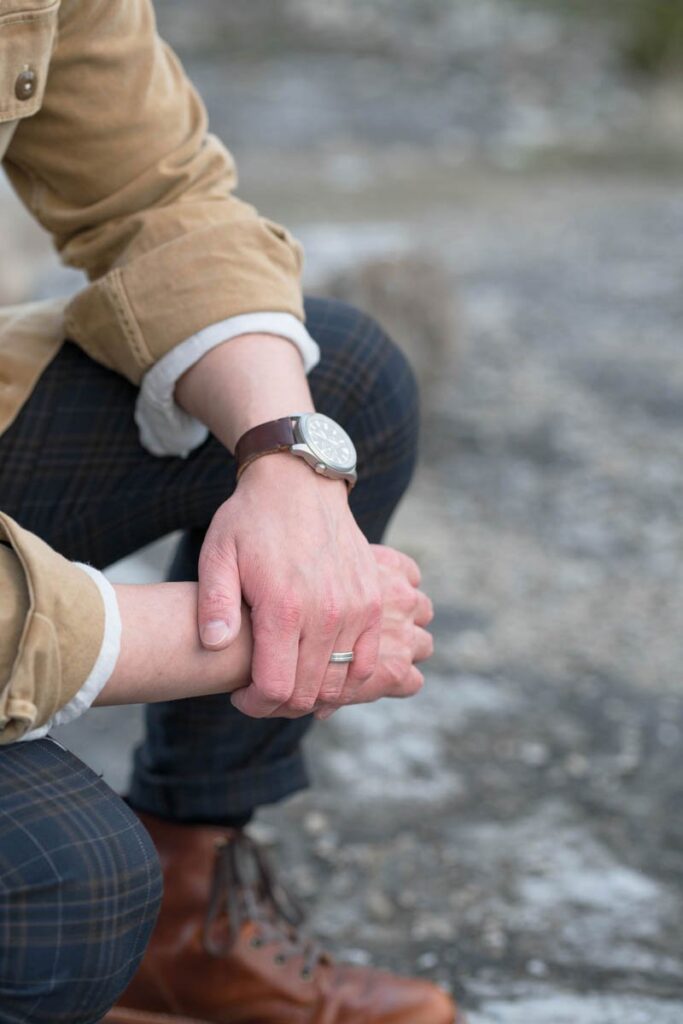 Groom crouches down clasping hands showing his wedding band and watch.