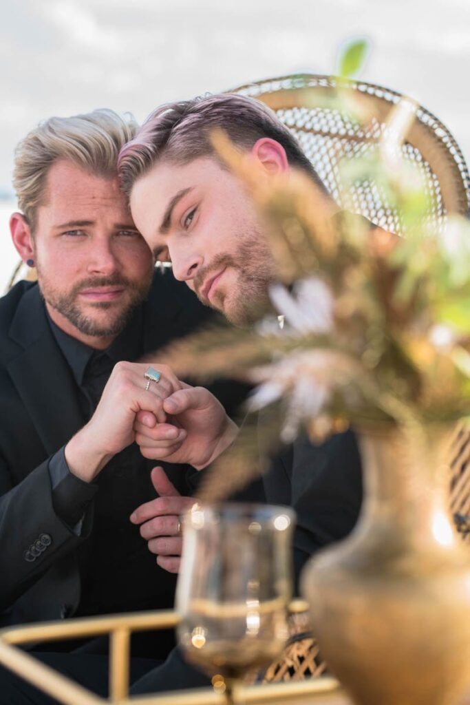 Grooms hold hands while sitting in wicker chairs.