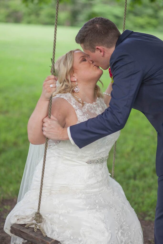 Bride and groom share a kiss while bride sits on wooden swing.