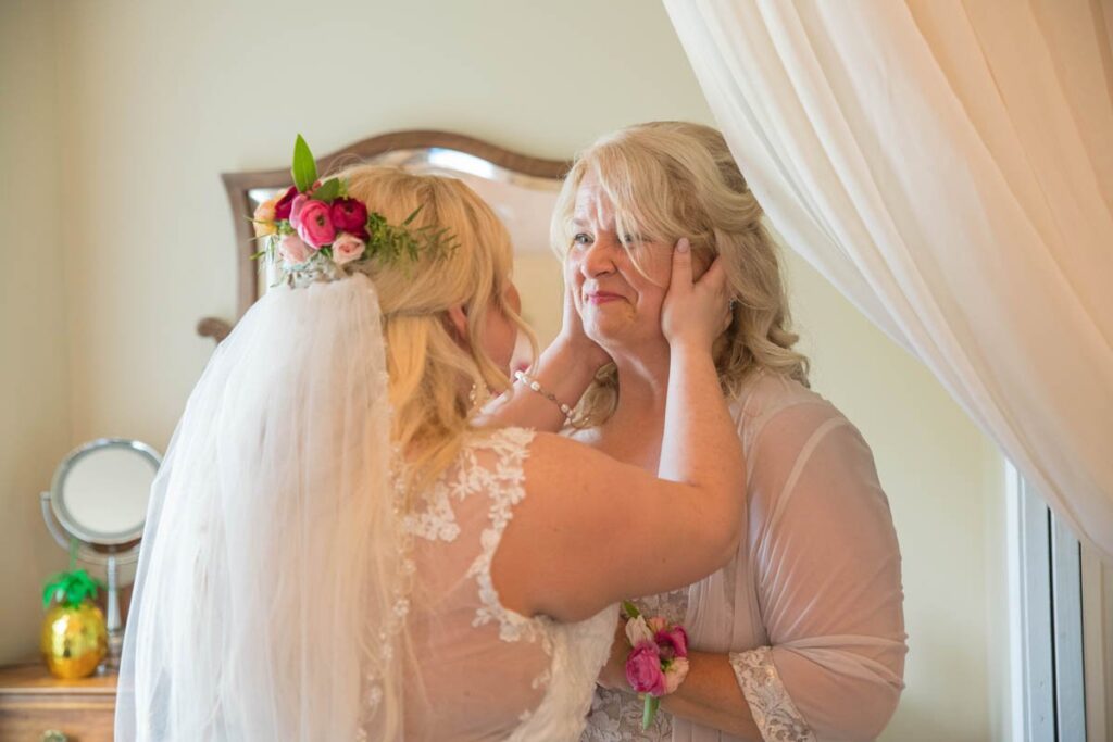 Bride holds her mother's face in her hands as mother smiles with tears in her eyes.