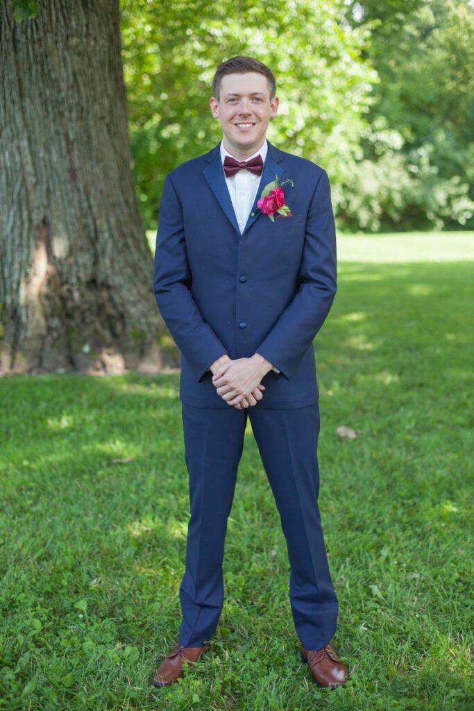 Groom stands smiling under large tree in grassy area.