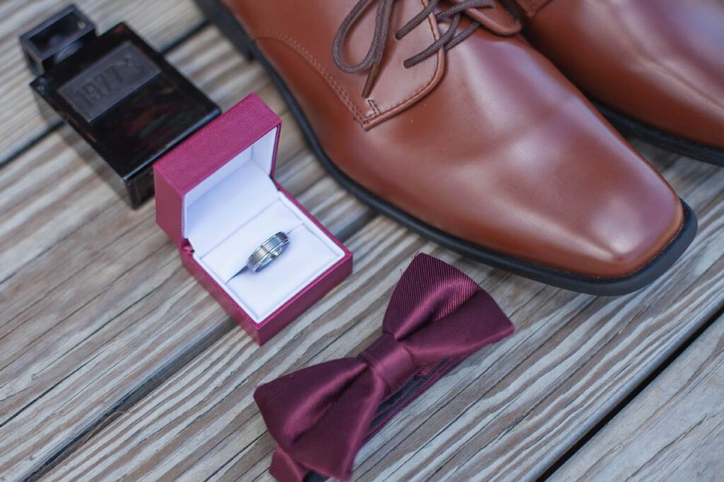 Groom's ring, maroon bowtie, shoes, and cologne on wooden boards.