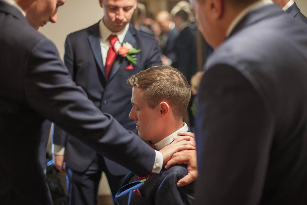 Groomsmen lay hands on sitting groom to pray for him.
