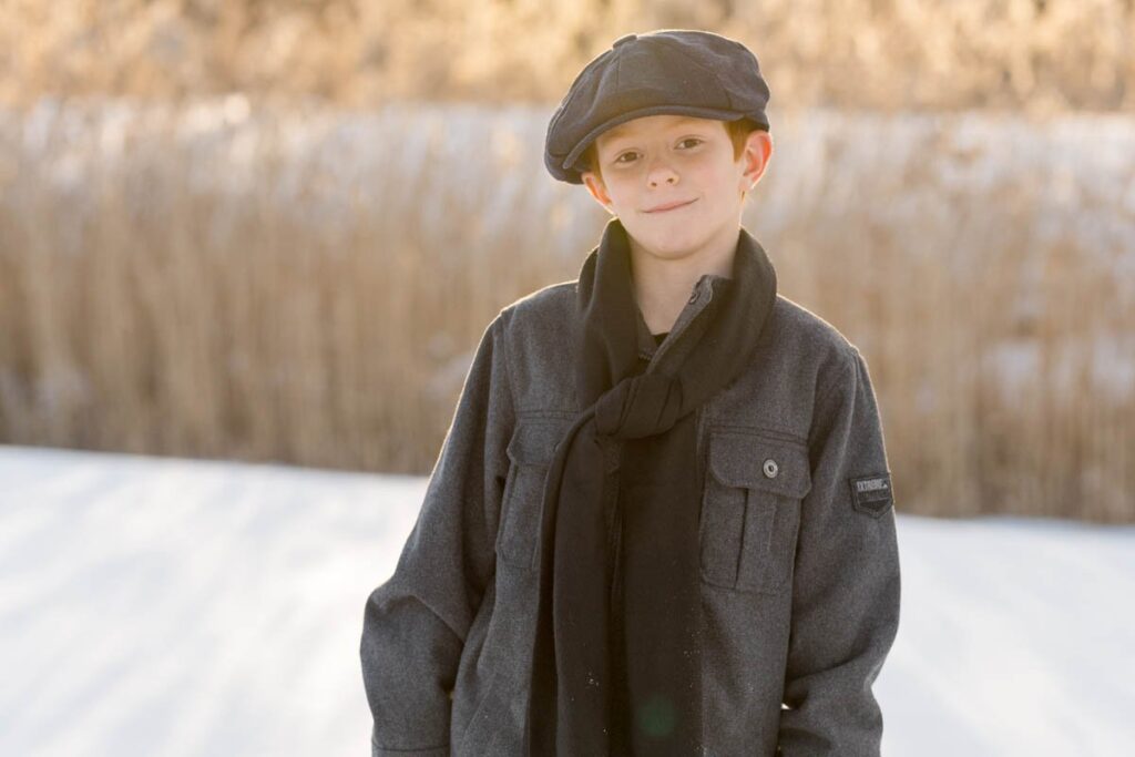 Boy in grey hat smirks with his hands in the pocket of his coat on a snowy day.