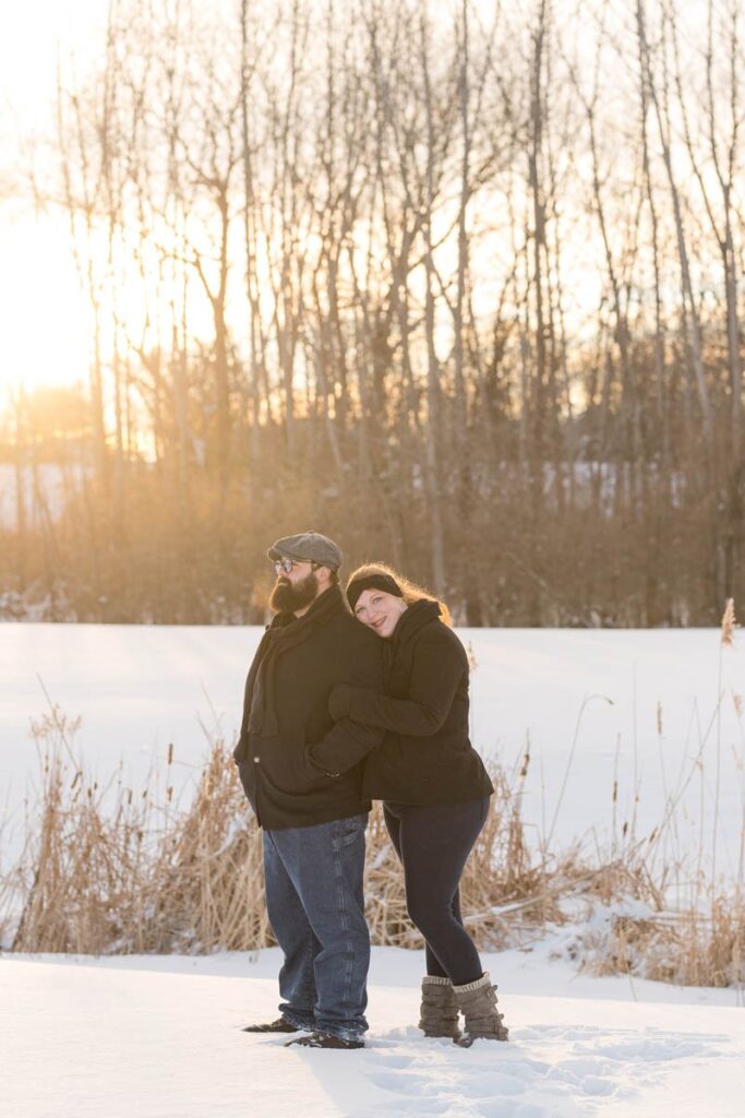 Couple smiles while standing together in the snow at sunset.