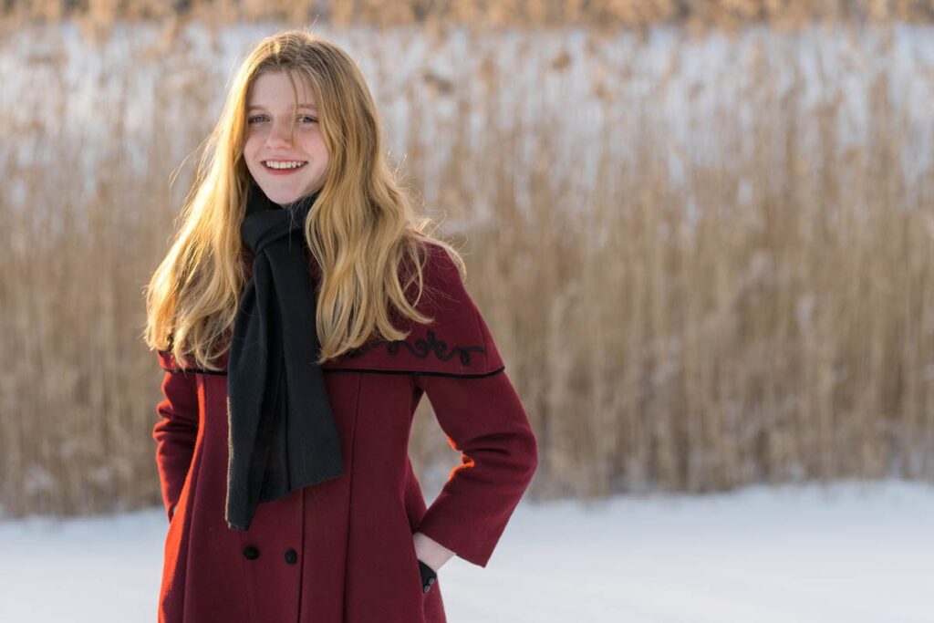 Girl wearing maroon coat with grey scarf smiles during photoshoot in the snow.