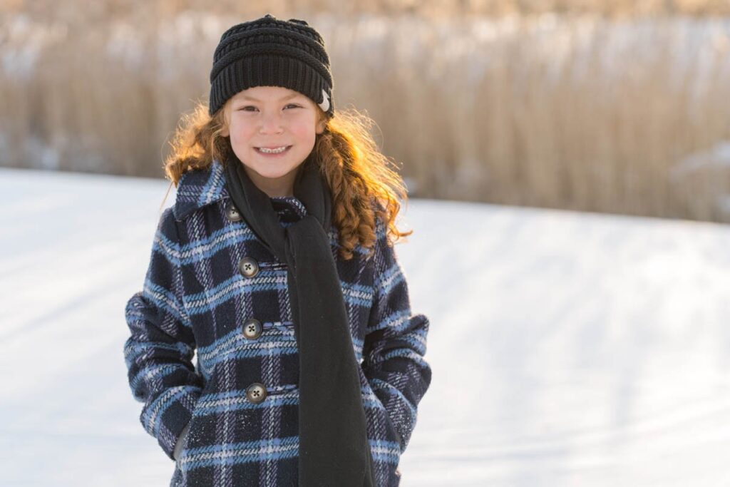 Little girl with red curly hair smiles as she stands in front of the sunset on a snowy day.