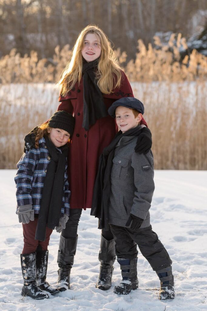 Siblings smile together in the snow with the sun setting behind them.