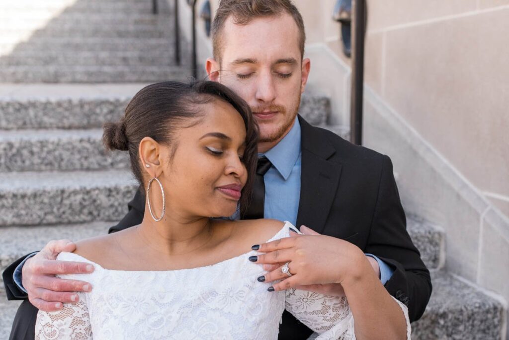 Bride leans back against groom while sitting on stone stairs with closed eyes.