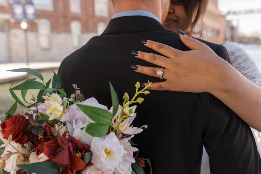 Bride and groom embrace and show off the bride's wedding ring with her bouquet in the foreground.