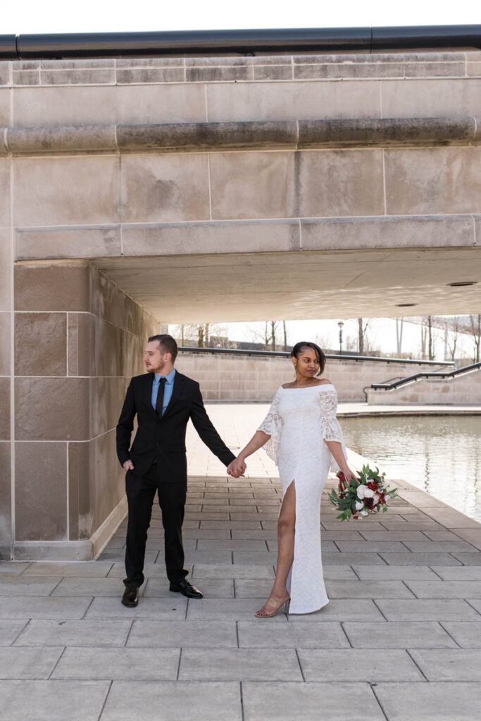 Bride and groom hold hands while standing under bridge looking opposite directions at their Indianapolis elopement.