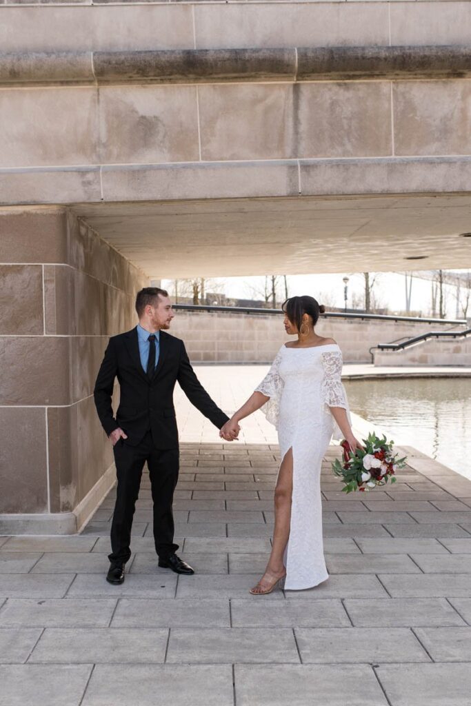 Bride and groom hold hands standing under a bridge looking at one another seriously.