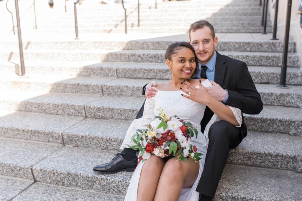 Bride and groom smile happily while sitting on stone stairs and bride shows off her ring.