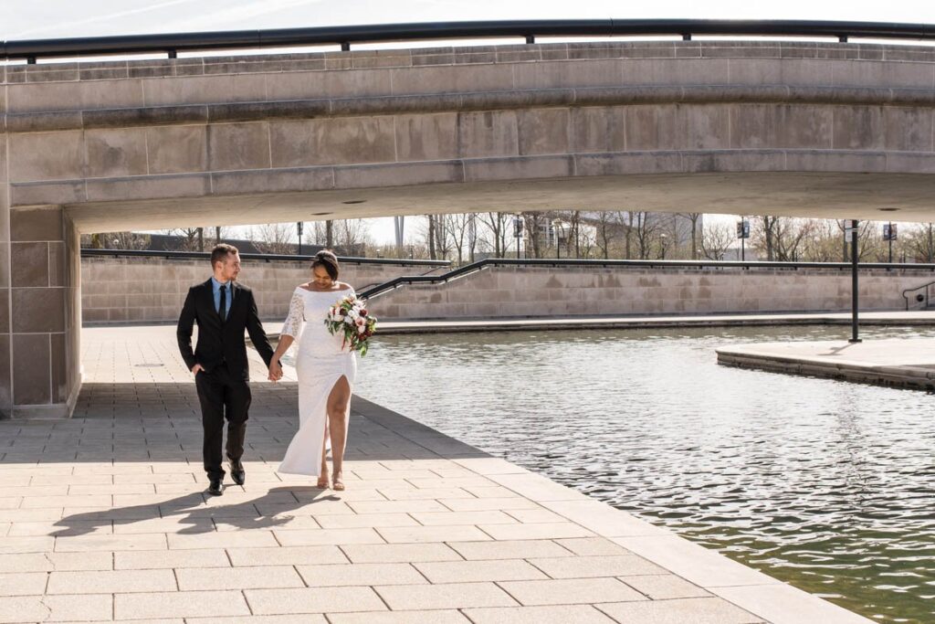 Bride and groom holding hands on the day of their Indianapolis elopement while walking alongside canal together.