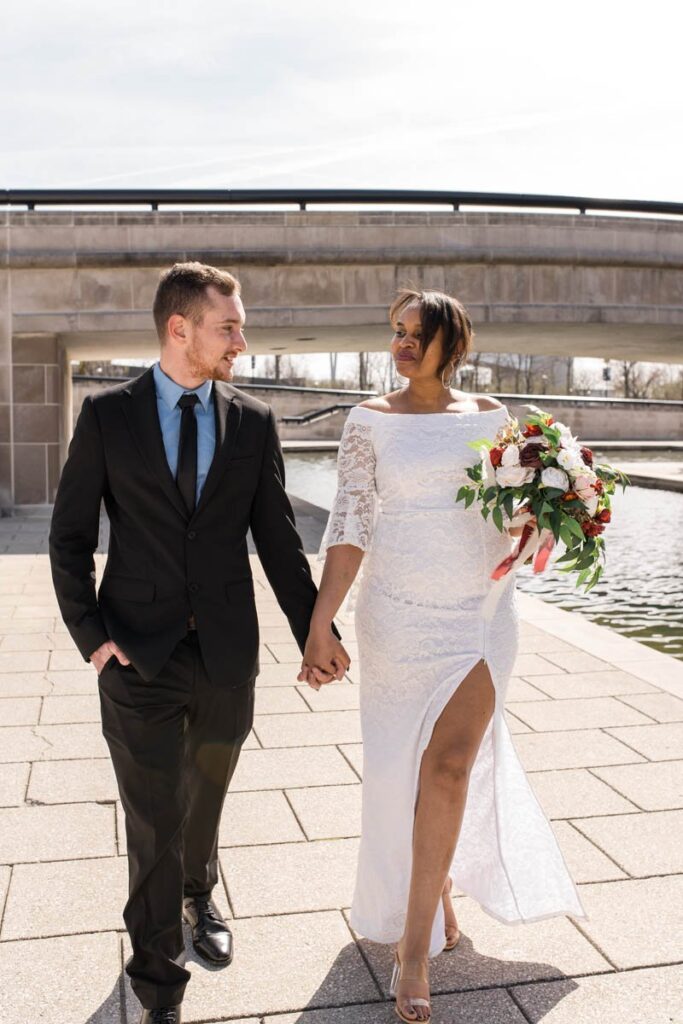 Bride and groom walk hand-in-hand smiling next to a canal with a bridge behind them.