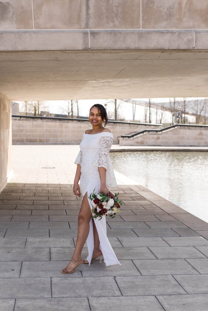 Bride smiles while holding the bouquet down at her side standing next to the canal.