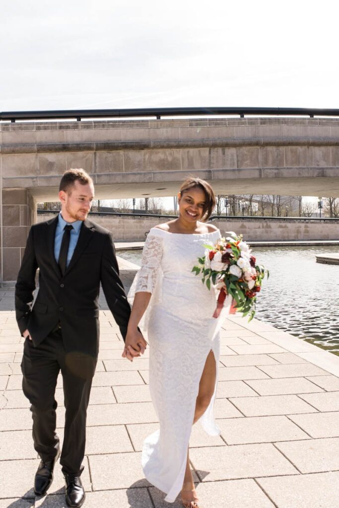 Bride smiles while holding grooms hand and walking along canal.