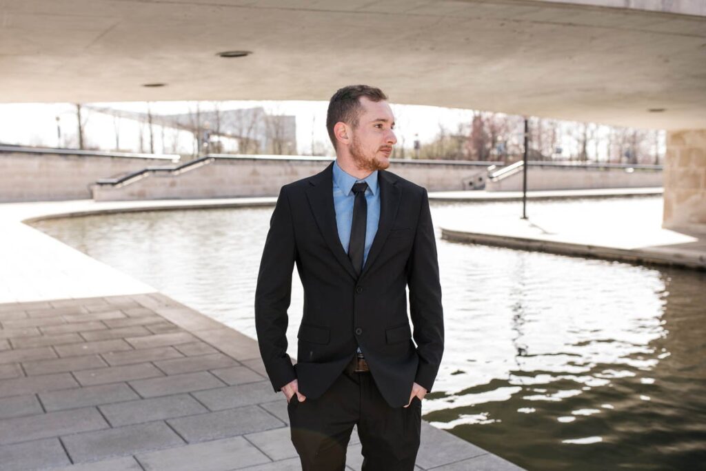 Groom stands under bridge next to water looking away to the right.