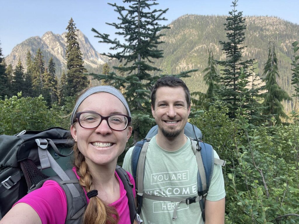 Leave No Trace Aware Photographers hiking with packs on and smiling with mountains and trees behind them.