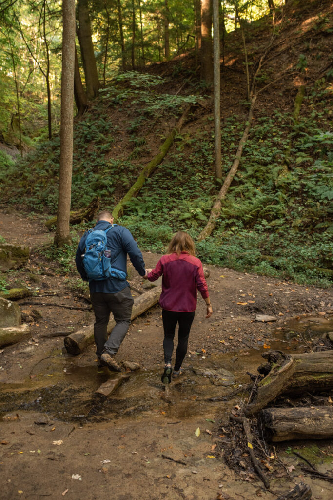Couple holds hands while hiking together through a small stream in the woods.