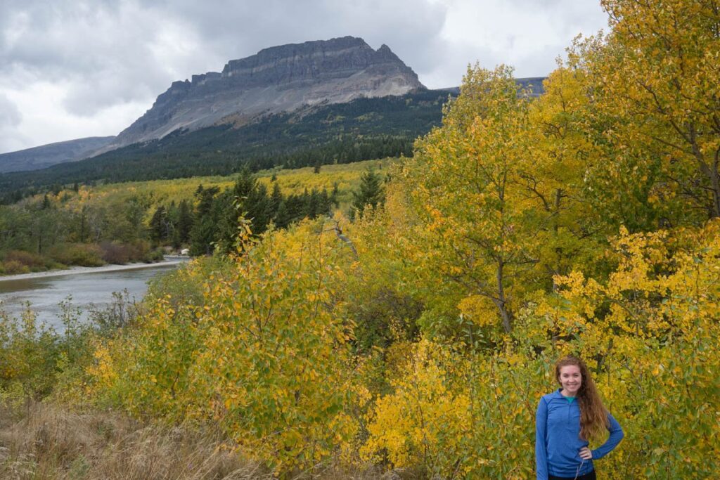 Woman in blue jacket is standing in front of trees with yellow leaves and large mountain is in the distance.