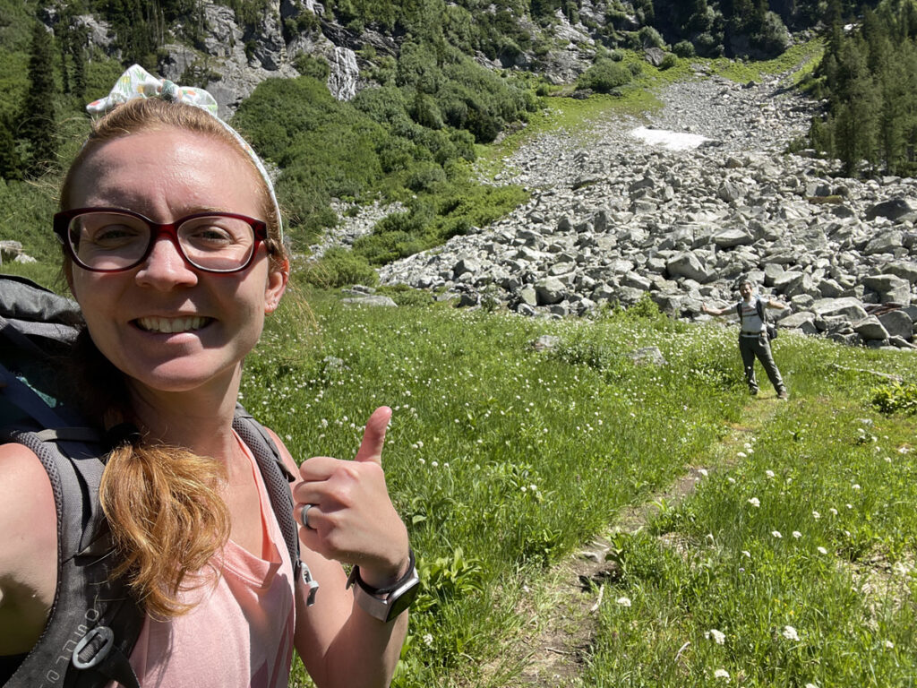 Two hikers give a thumbs up which walking on a small trail among wildflowers in North Cascades National Park.