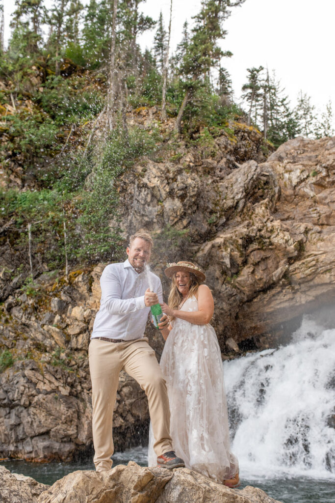 Bride and groom pop sparkling water after they exchange vows in front of a waterfall at Glacier National Park.